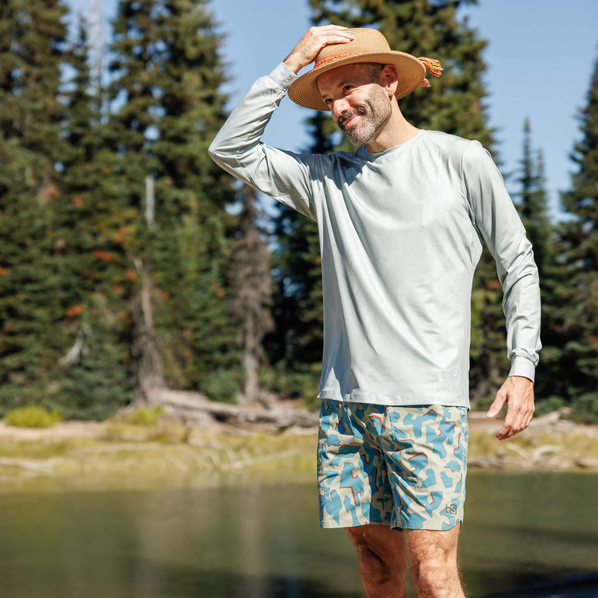 Man standing near an alpine lake with a snow-capped mountain in the background wearing the BlackStrap Men's Brackish Long Sleeve shirt #color_corally spruce