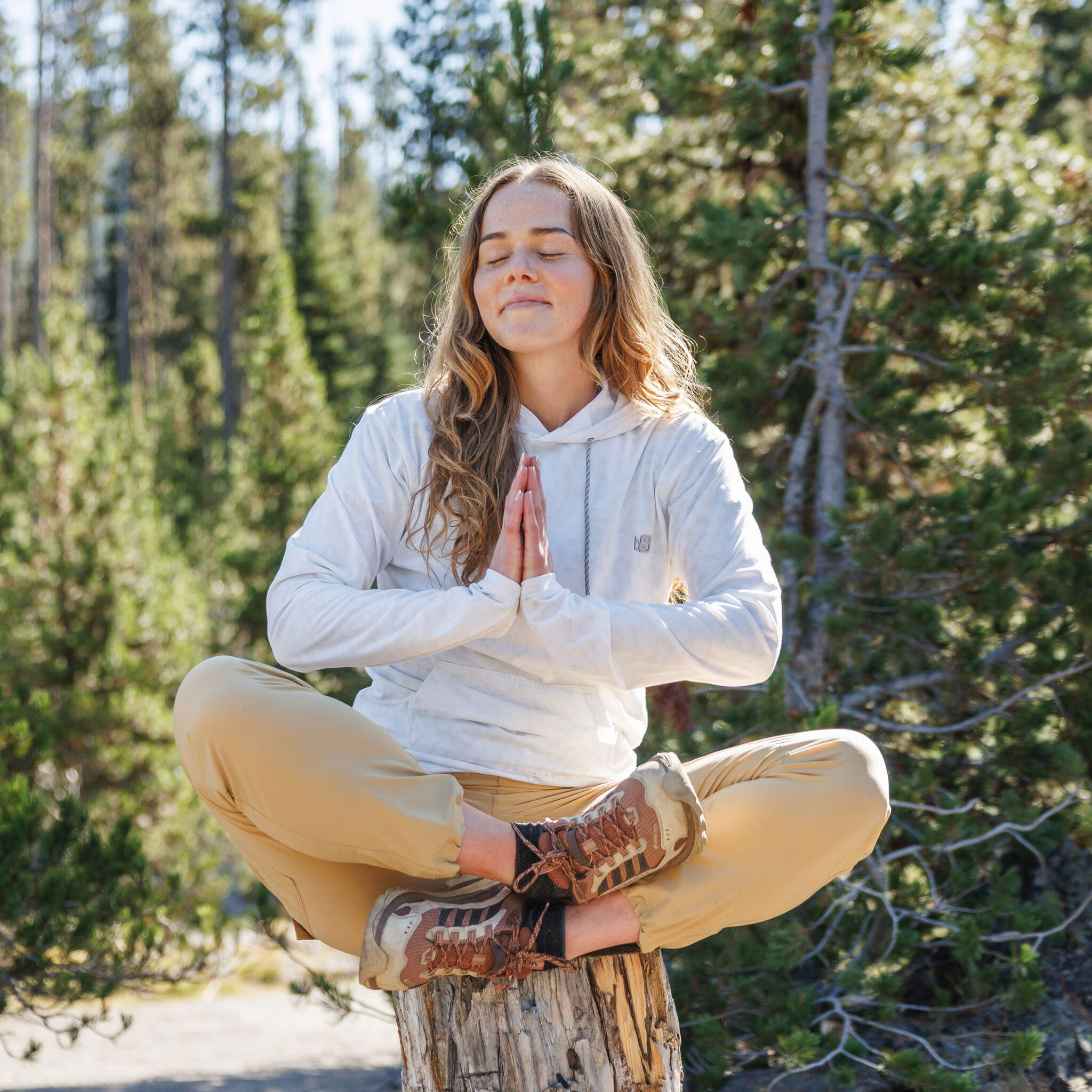 Woman meditating on a tree stump in a pine forest wearing the BlackStrap Women's Aspect Hoodie Long Sleeve #color_ripple ash