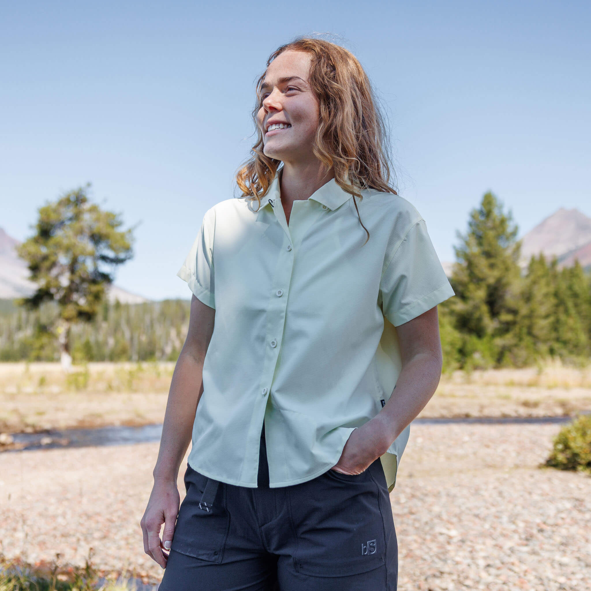 Woman smiling outdoors wearing a Orion short-sleeve BlackStrap camp shirt with utility pants, with pine trees and mountains in the background.