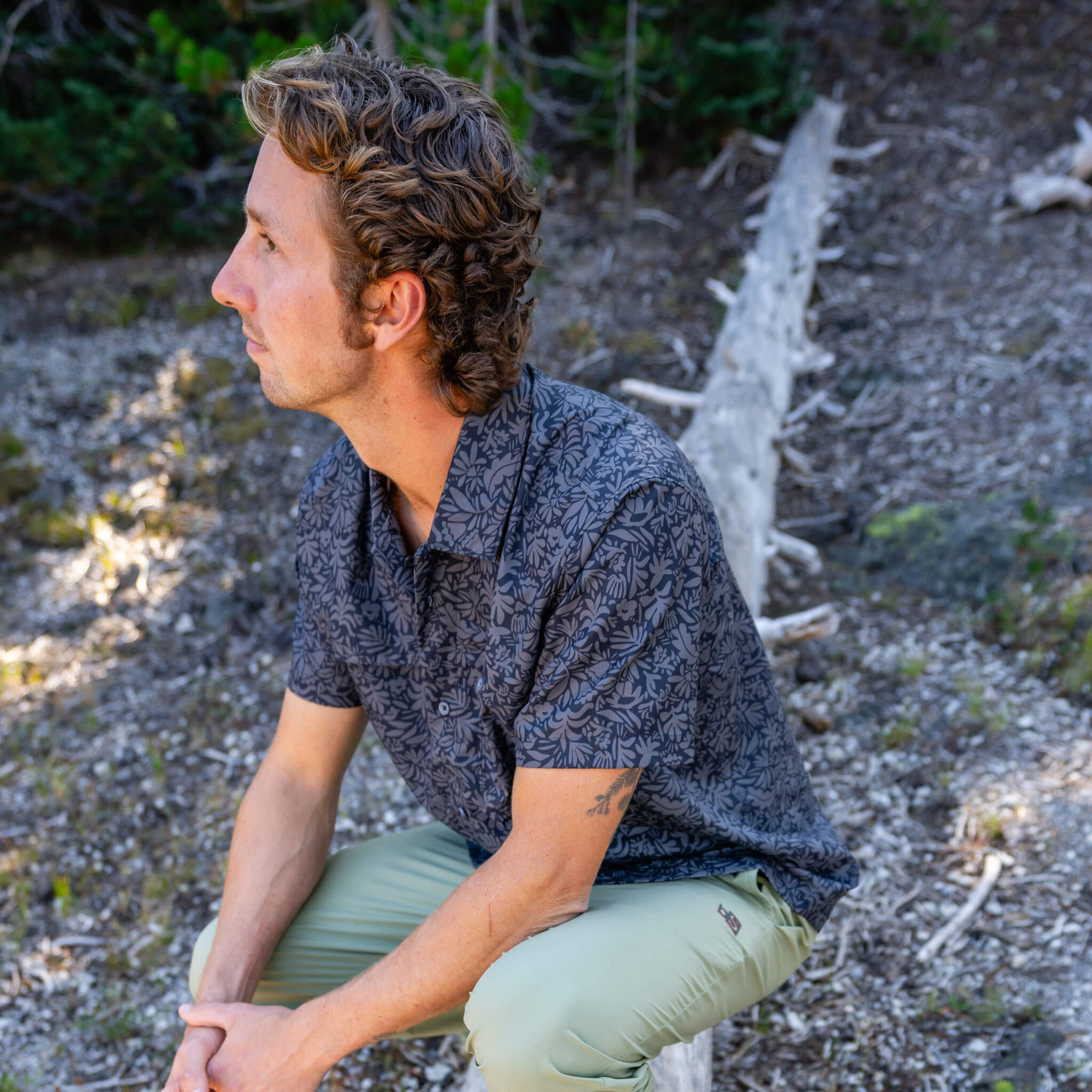 Man sitting outdoors in profile wearing a florican deschutes short-sleeve BlackStrap camp shirt and approach pants, with a rocky forest floor and fallen trees behind him #color_florican smoldered