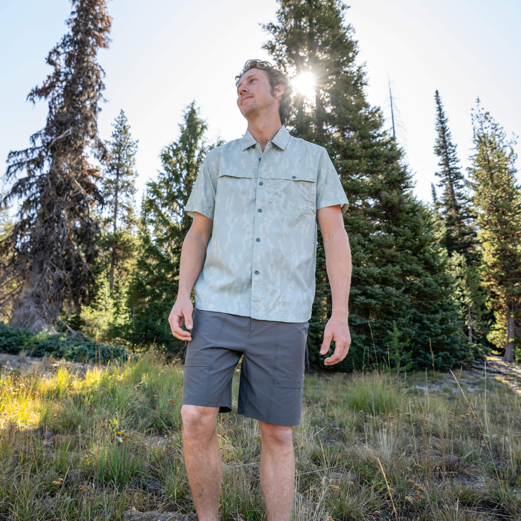 Man standing in an alpine meadow looking upward, wearing a Corally Spruce short-sleeve BlackStrap camp shirt with chest pockets and utility shorts, with pine trees and sunlight behind him. #color_corally spruce