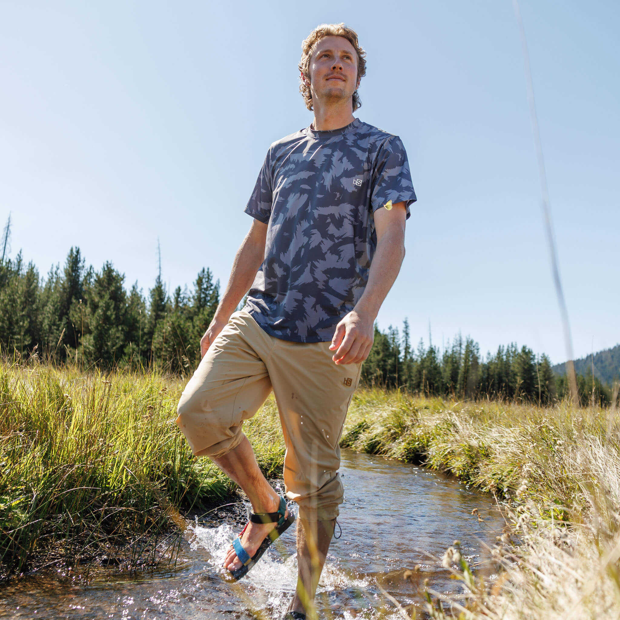 Man wading through a mountain meadow stream wearing the BlackStrap Men's Voyage Tee #color_slash smoldered