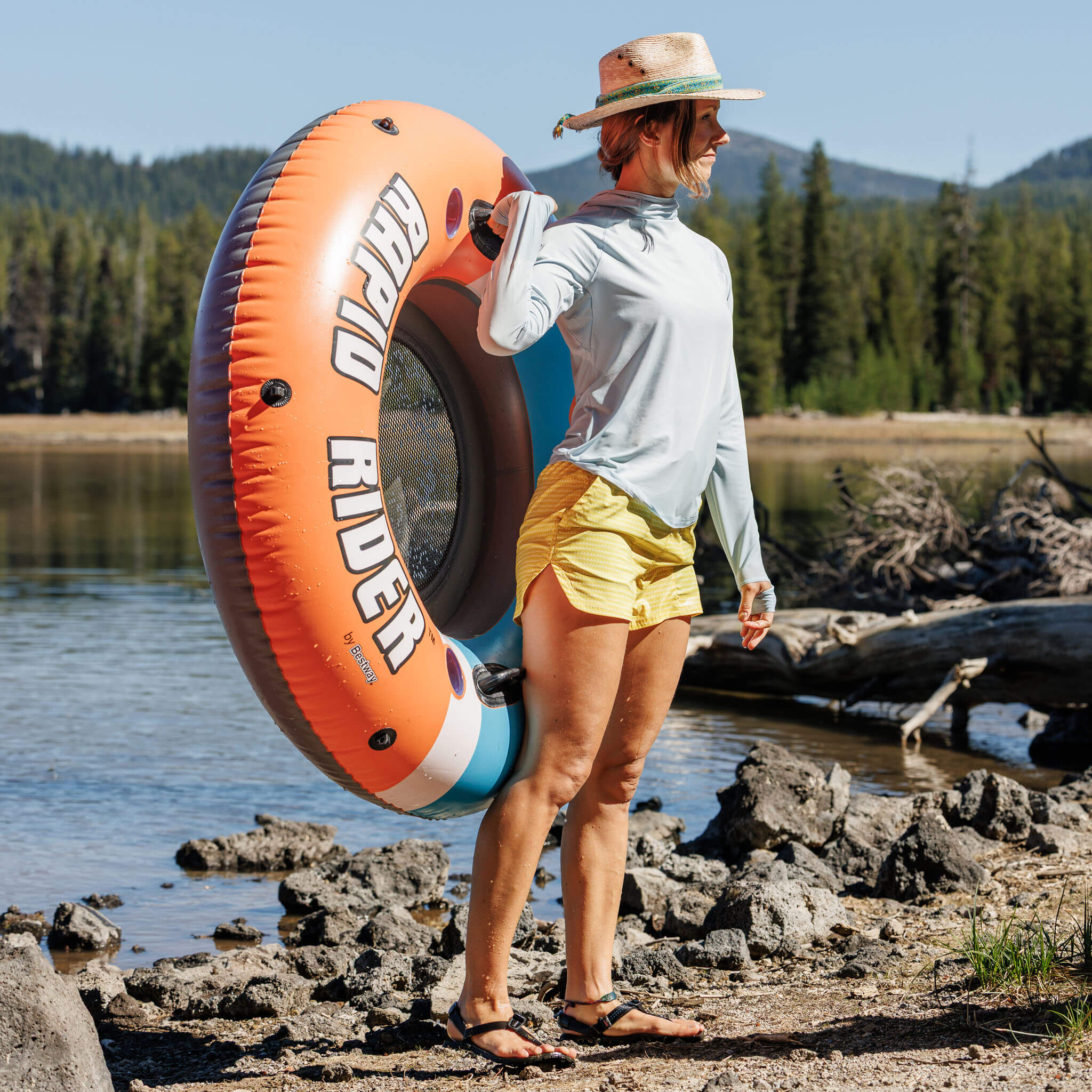 Woman carrying a river tube along a rocky lakeshore wearing the BlackStrap Women's Brackish Long Sleeve Hoodie #color_stratus