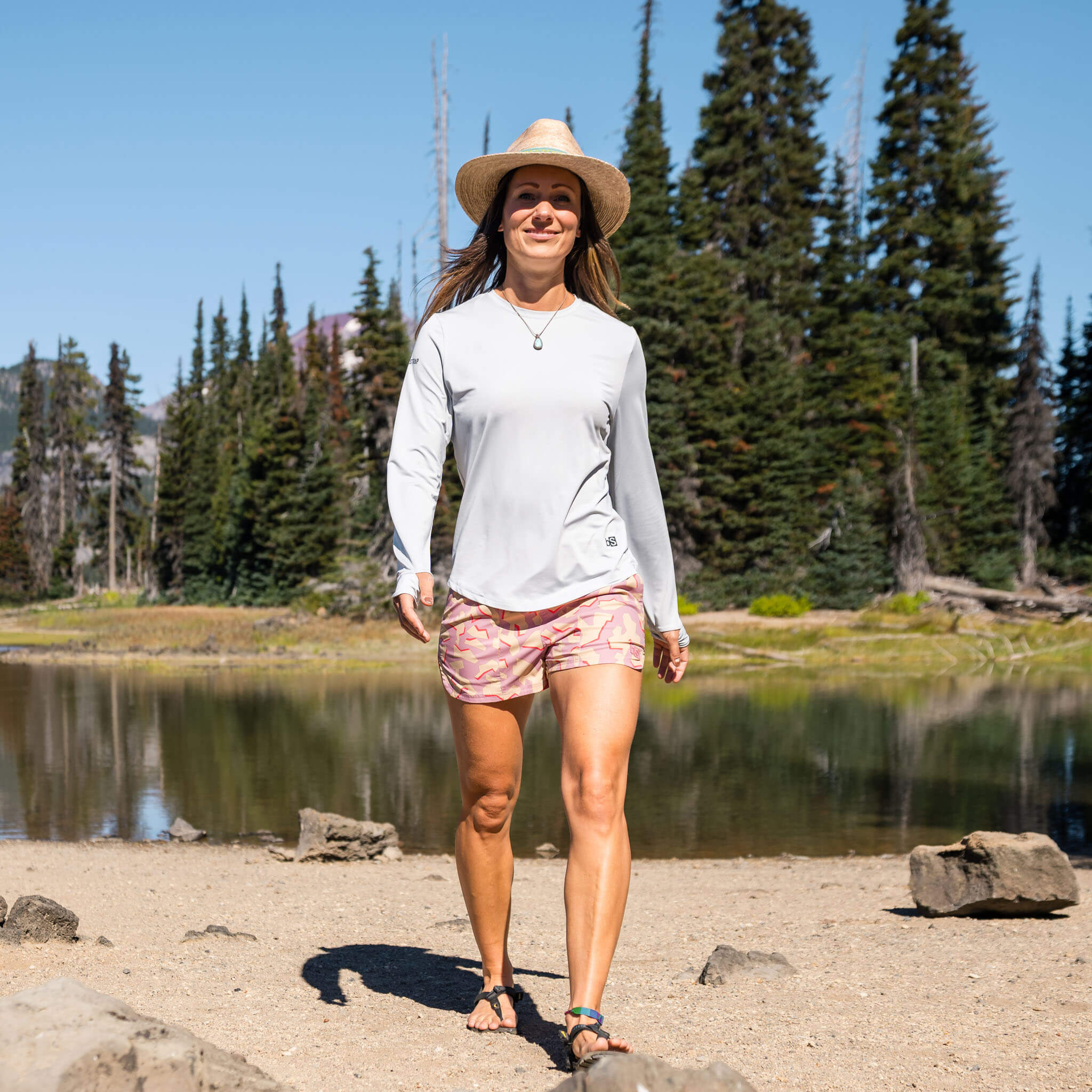 Woman walking along a sandy alpine lakeshore wearing the BlackStrap Women's Brackish Long Sleeve shirt  #color_stratus